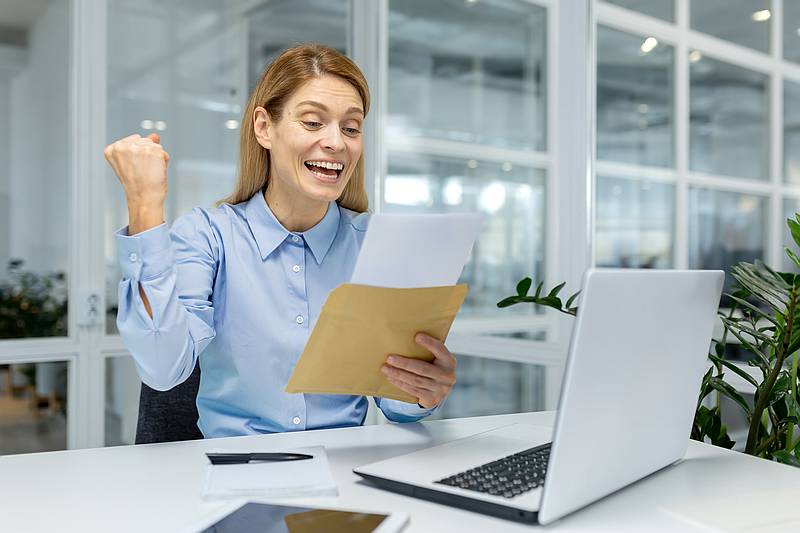 A joyful female office worker celebrates success with an envelope in a modern workspace, evoking feelings of achievement and happiness.