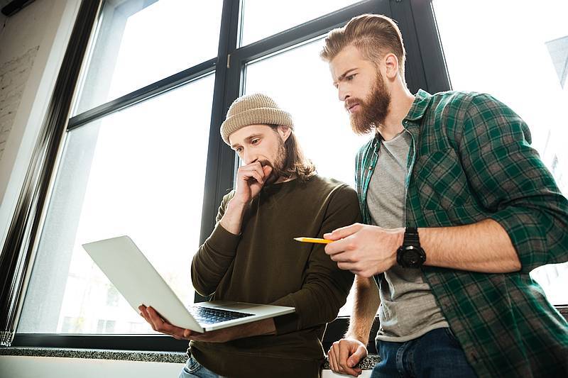 Image of young concentrated men colleagues in office using laptop computer. Looking aside.